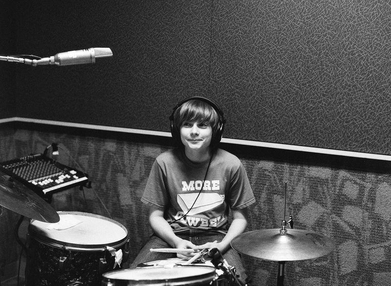 Spencer sitting at James Gadsons drums, wearing a More Cowbell t-shirt, photo by Autumn de Wilde