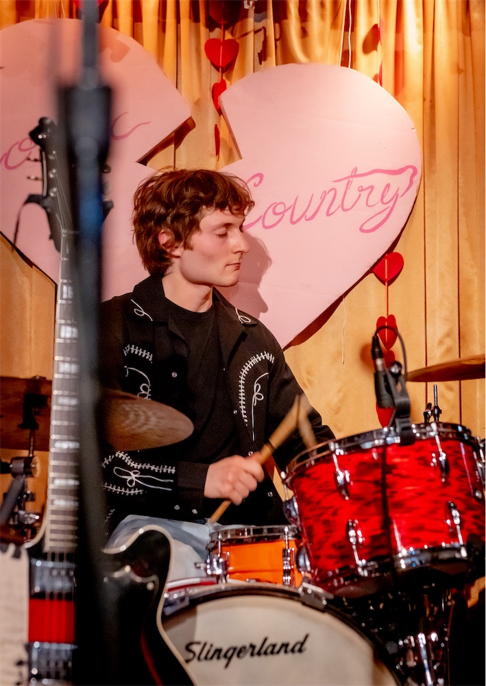 Spencer Tweedy playing a red onyx Slingerland drum kit, eyes closed, in front of yellow curtains.