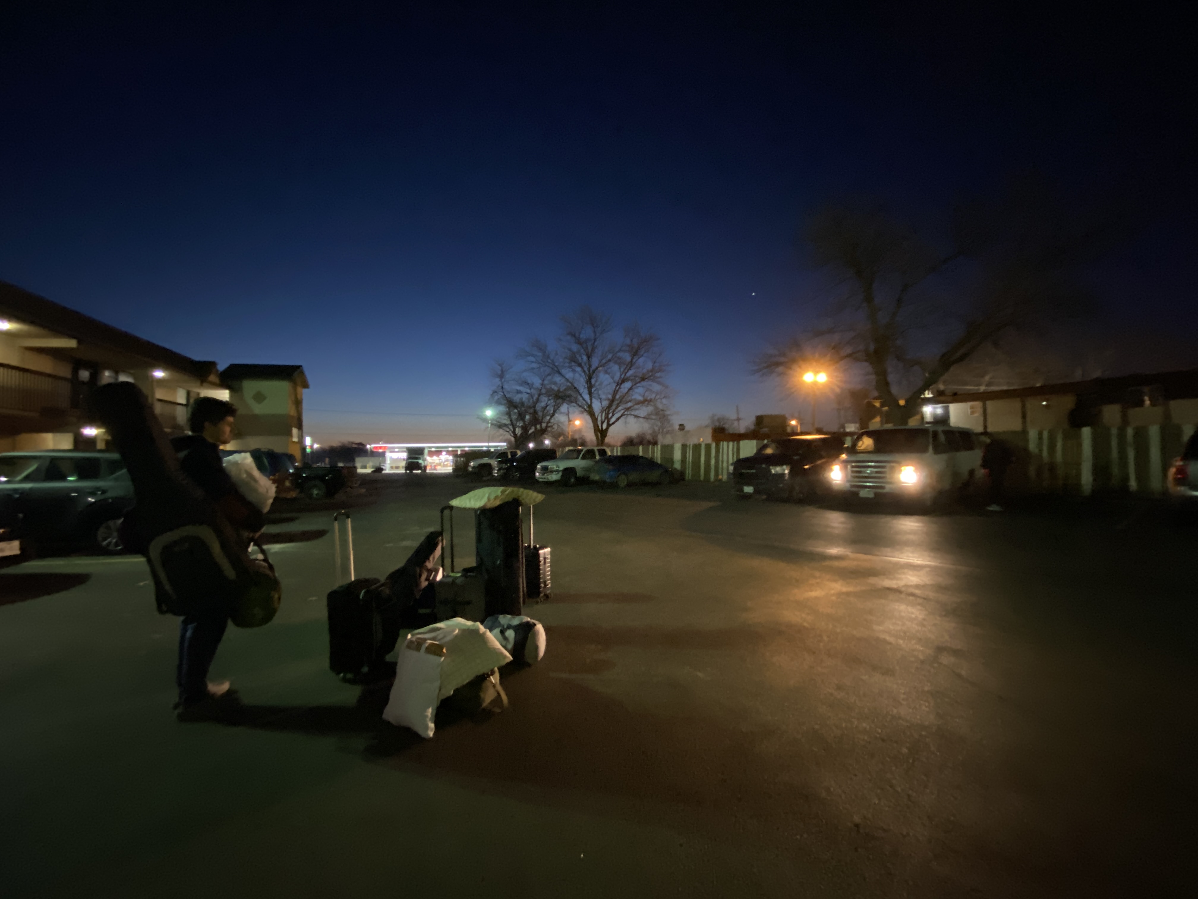 Lane stands with the band’s bags in the parking lot of a motel in Tulsa, Oklahoma, at dawn, illuminated by our van’s headlights.