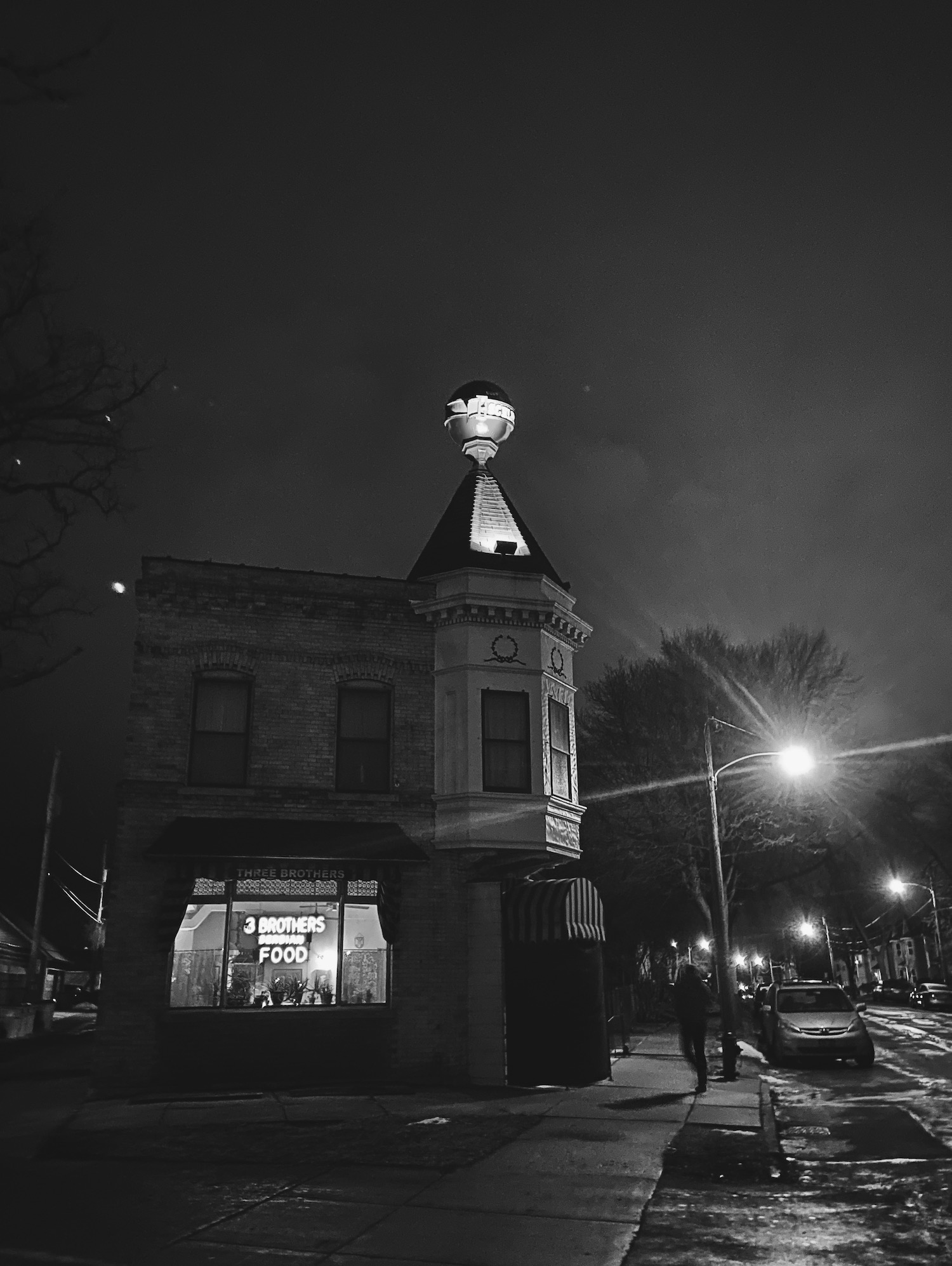A black-and-white photo of a restaurant with a neon sign that reads 3 Brothers Serbian Food in its window, with an illuminated Schlitz Beer globe on its roof, on a quiet Milwaukee street.
